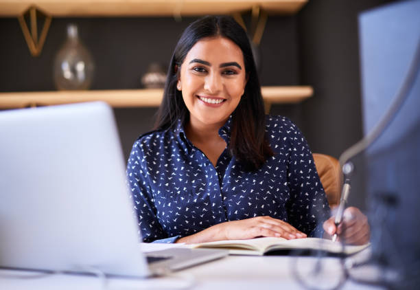 istock Happy indian businesswoman writing in a notebook while working alone on a laptop in an office. One female only making notes in her diary and planning her schedule while working at her desk 1396321416