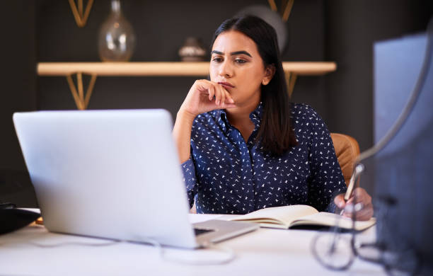 Focused indian businesswoman writing in a notebook while working alone on a laptop in an office. One female only thinking before making notes in her diary and planning online while working at her desk