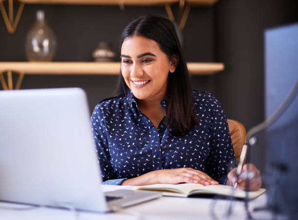 istock Young indian businesswoman writing in a notebook while working alone on a laptop in an office. One female only making notes in her diary and planning online while working at her desk 1396321421