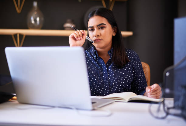 istock Focused indian businesswoman writing in a notebook while working alone on a laptop in an office. One female only thinking before making notes in her diary and planning online while working at her desk 1396321429