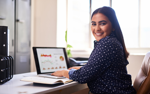 Happy young indian businesswoman working alone on a laptop in an office. One female only analysing graphs and statistics on a laptop at her desk