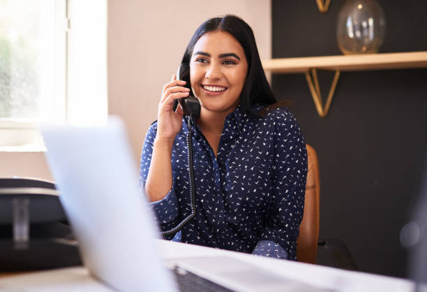 giovane donna d'affari indiana che parla al telefono in un ufficio da sola. una donna effettua solo una chiamata mentre lavora come receptionist presso una reception. consulenza di amministratori e segretari e trasferimento di chiamate da un help desk in u - adult middle eastern ethnicity asian ethnicity cheerful foto e immagini stock