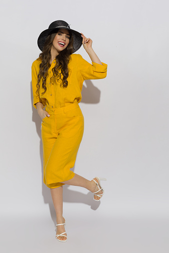 Cheerful young woman wearing a linen shirt, shorts, black hat and high heels poses on a one leg. Full length studio shot.