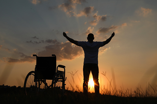 Man raising hands up to sky near wheelchair at sunset, back view. Healing miracle