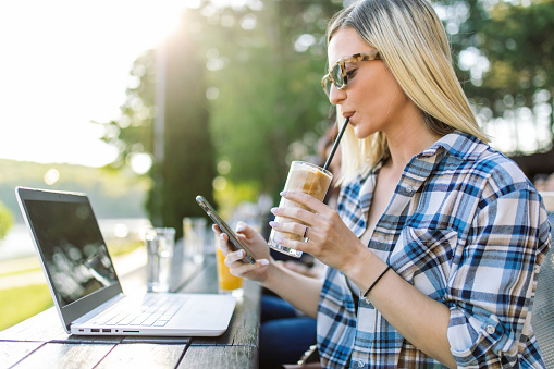Young blonde woman working on laptop on her vacation. She is enjoying good smell of coffee frappe and freedom to combine work and travel. She is wearing business casual clothes and looks beautiful and carefree