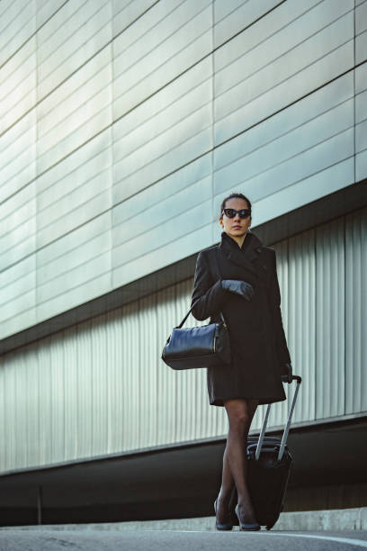 istock Young elegant woman walks down the sidewalk next to an office building and pulls small luggage on wheels 1396353441