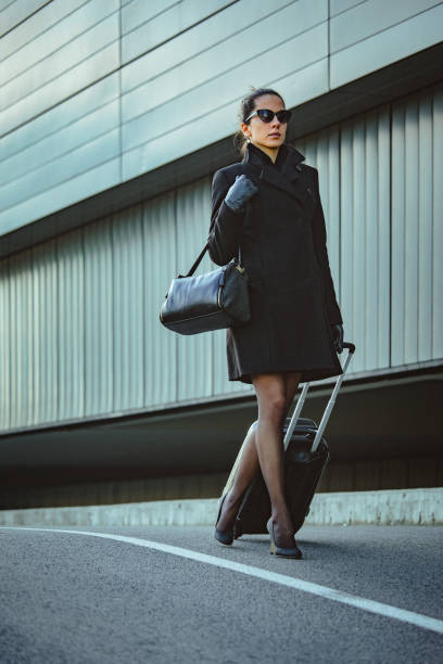 istock Young elegant woman walks down the sidewalk next to an office building and pulls small luggage on wheels 1396353461