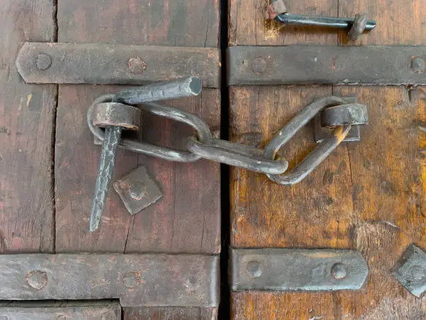 close-up of old wooden doors from the medieval, middle ages. Forged iron braces holding wood together. Chain links going from one side to the other with a bent iron spike in hasp to look doors together.