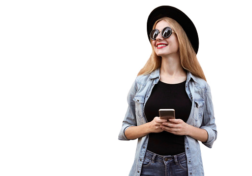 Portrait of happy smiling young woman with smartphone looking away wearing black round hat, jeans jacket isolated on white background