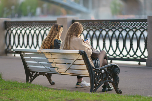 Moscow, Russia - May 6, 2022: Young women sitting on the bench in the public Gorky park. They are resting in the sunny warm day. They using smartphone, looking at the screens. Lifestyle concept. Backs