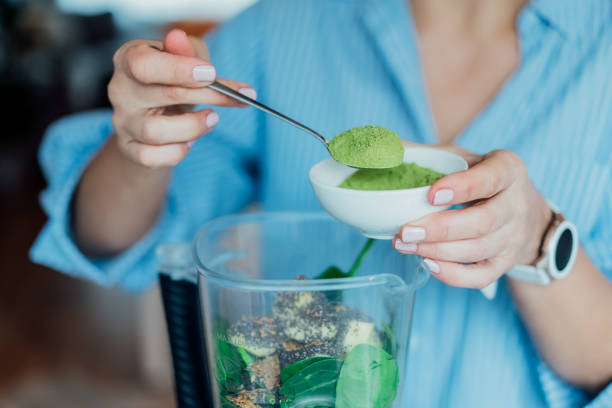 feche a mulher adicionando pó verde de grama de trigo durante fazer smoothie na cozinha. suplemento de superalimentos. dieta vegana desintoxicação saudável. alimentação saudável, programa de perda de peso. foco seletivo - só uma mulher madura - fotografias e filmes do acervo