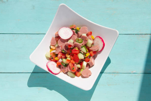 A traditional Romanian lunch with plates of fresh vegetables - spring onions, tomatoes, cucumbers. There is fresh meat including salami, sausage and slanina - cubes of pork fat. There are hard boiled eggs and a loaf of fresh bread. All of this is presented on a white wooden surface.