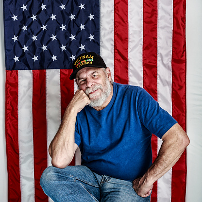 An authentic, real person USA Vietnam War military veteran - with his right hand fist on his chin and his left hand fist on his hip - is relaxed. He is sitting in front of an American flag background and looking at the camera with a friendly, confident facial expression.