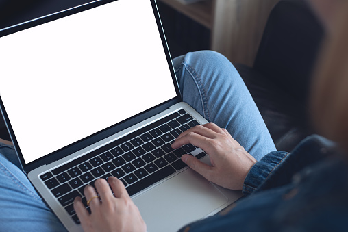 Laptop computer mockup, young casual business woman sitting on sofa, working on blank screen laptop computer at home, close up, over shoulder view