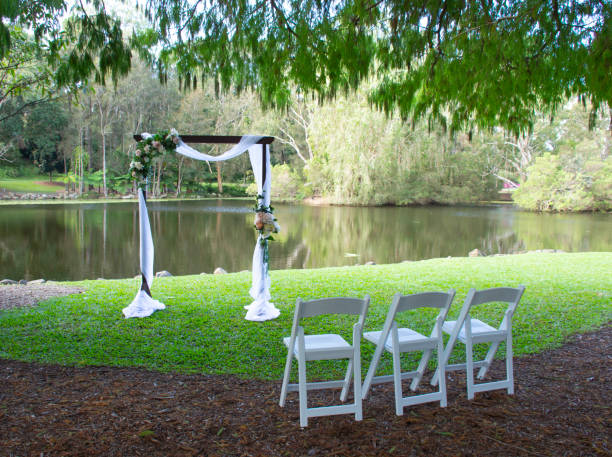 istock Three white chairs facing a rectangle arch ready for a wedding.  Brown bark and green grass under a leafy tree.  Reflections on the pond in the background. Gold Coast Queensland Australia 1396388070