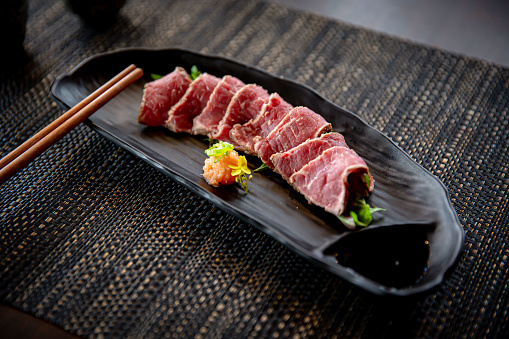 Beef tataki appetizer in the traditional Japanese ramen restaurant, with a black plate on a black mat in a black scene background