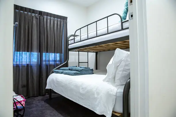 Bright white modern interior with dual-layered bunk bed, grey curtain and travel suitcase in accommodation hostel hotel holiday stay, with bedding, pillows, green blanket