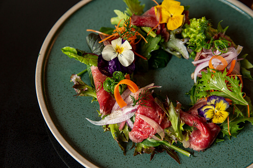 Tartare beef flower circle salad in the fine dining Japanese restaurant, with a green round plate in black scene reflection table background, raw beef, onion, salad, carrot, flower decorations