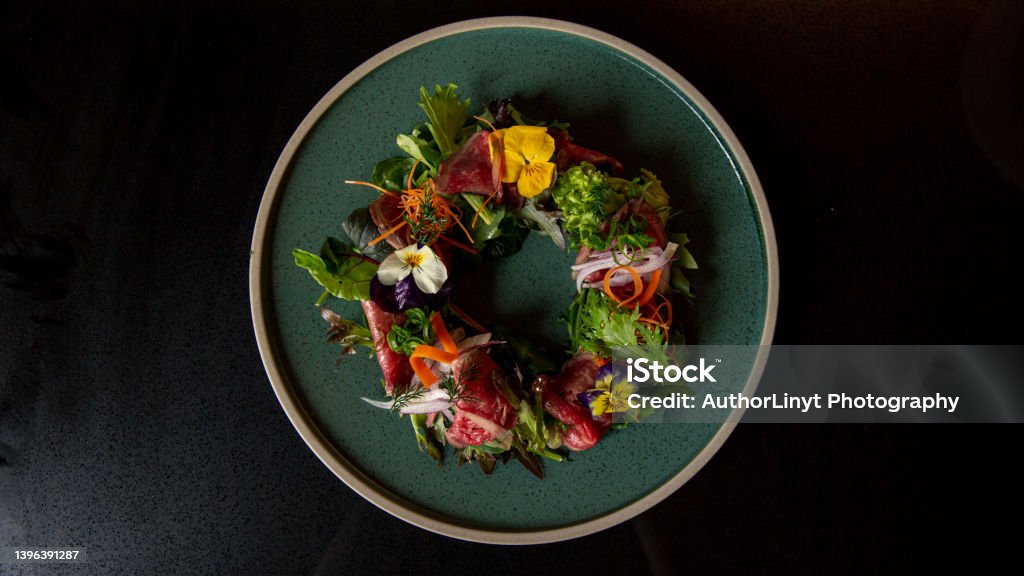 Top view of Tartare beef flower circle salad in the fine dining Japanese restaurant Top view of Tartare beef flower circle salad in the fine dining Japanese restaurant, with a green round plate in black scene reflection table background, raw beef, onion, salad, carrot, flowers Crockery Stock Photo