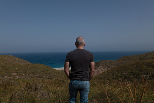 Rear view of adult man standing on hill looking at sea against blue sky