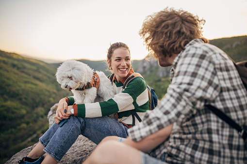 Man and woman, couple of hikers and their pet dog sitting on a rock high on mountain in sunset.