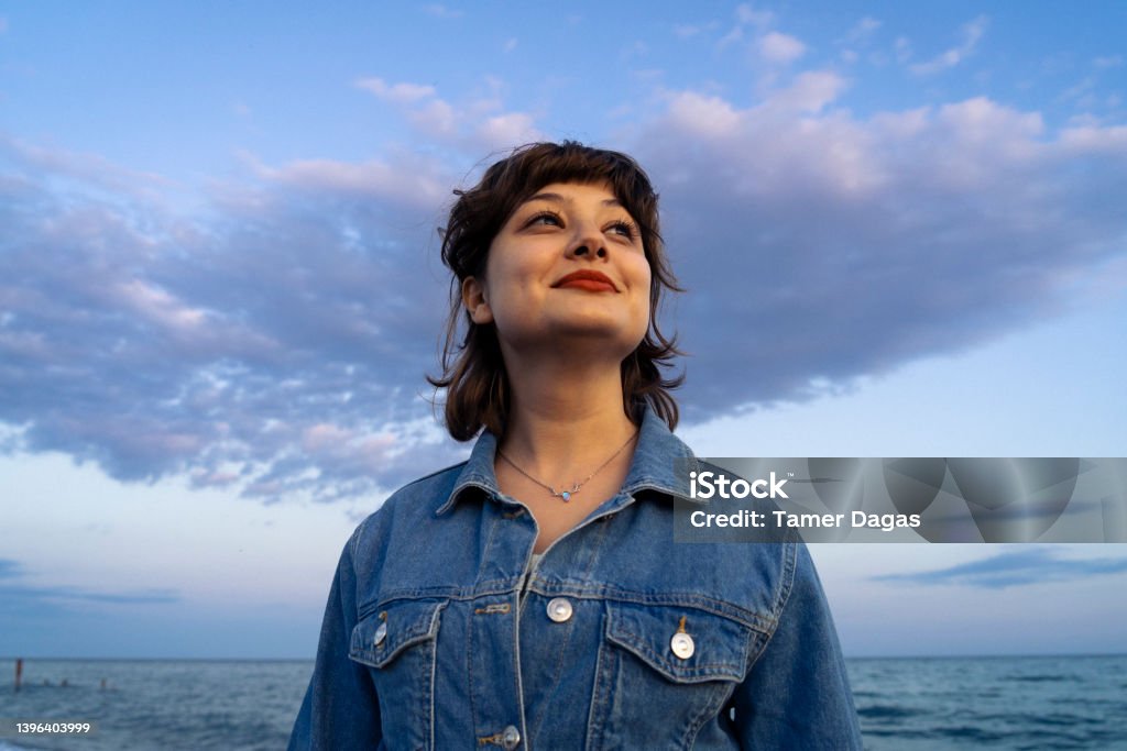 Look to the future with hope! Photograph of a beautiful young woman wearing a denim jacket and smiling towards the sun, taken in the sweet pastel colors of the sunset. The Way Forward Stock Photo
