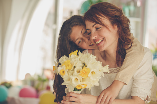Copy space shot of a cheerful mid adult woman smiling after her young daughter, who is embracing her from behind, surprised her with a bouquet of yellow daffodils for Mother's day.