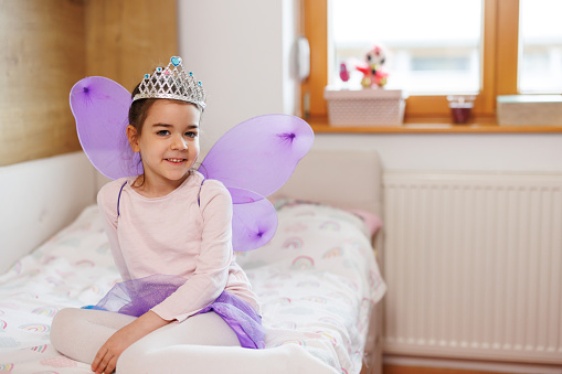 Portrait of a cute happy little girl wearing a princess crown and purple fairy costume, sitting on bed and looking at camera at home