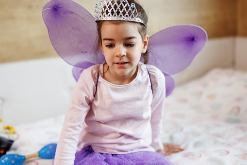 Close-up shot of cute little girl with purple fairy costume and silver diadem sitting on bed in her room