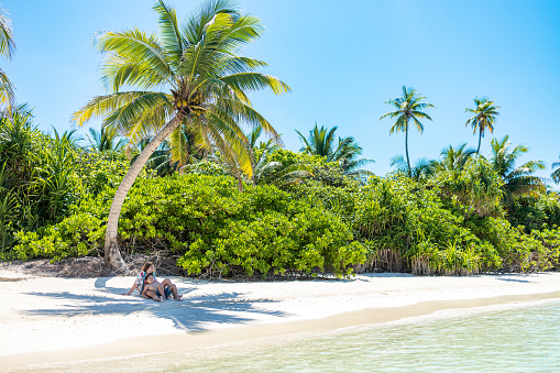 Woman and child lying in the shade of a palm tree on a white sand beach