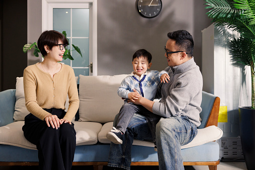 Young asian parents, indoors during the day, hold their son and play on the sofa.