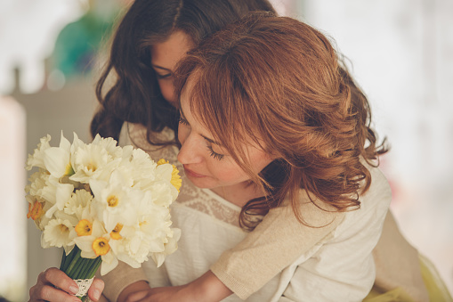 Happy mid adult woman smelling the beautiful bouquet of yellow daffodils she got from her affectionate daughter, for Mother's day, who is embracing her from behind.