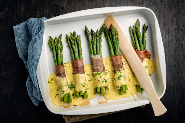 Asparagus Wrapped With Parma Ham and Hollandaise Sauce. Overhead view of bunch of green asparagus wrapped in parma ham with a hollandaise sauce. The last few woody centimetres of the asparagus stalk have been removed and the stalks have been cooked gently in a mix of boiling water, lemon juice, and salt for between 6-8 minutes depending on the thickness of the stalk. Then, depending on the size of the stalk 5 or 6 are wrapped with parma ham and baked in the oven for 10-12 minutes with a hollandaise sauce poured over. Colour, horizontal format with some copy space. Served as a vegetable accompaniment or on its own for a light lunch. hollandaise sauce stock pictures, royalty-free photos & images
