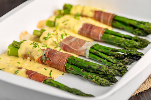 Bunches of green asparagus wrapped in parma ham with a hollandaise sauce. The last few woody centimetres of the asparagus stalk have been removed and the stalks have been cooked gently in a mix of boiling water, lemon juice, and salt for between 6-8 minutes depending on the thickness of the stalk. Then, depending on the size of the stalk 5 or 6 are wrapped with parma ham and baked in the oven for 10-12 minutes with a hollandaise sauce poured over. Colour, horizontal format with some copy space. Served as a vegetable accompaniment or on its own for a light lunch.