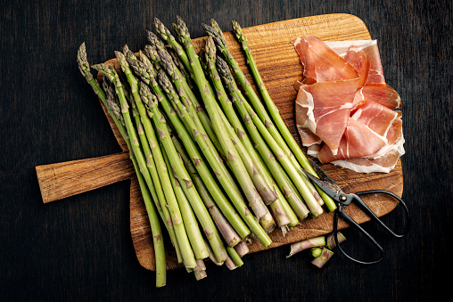 Overhead view of bunch of green asparagus wrapped in parma ham. The last few woody centimetres of the asparagus stalk have been removed and the stalks have been cooked gently in a mix of boiling water, lemon juice, and salt for between 6-8 minutes depending on the thickness of the stalk. Then, depending on the size of the stalk 5 or 6 are wrapped with parma ham and baked in the oven for 10-12 minutes. Colour, horizontal format with some copy space. Served as vegetable accompaniment or on its own for a light lunch.