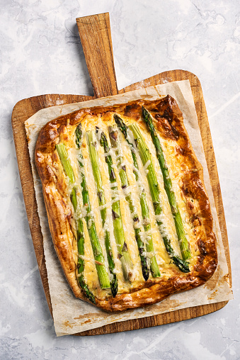 Overhead view of an asparagus and cheese tart. A shop bought short crust pastry dough has been used as a base for the tart. Recipe; the pastry base has been rolled out and an area about 2cm from the edge has been scored all around and brushed with egg, the rest of the base has been pricked with a fork and part cooked for 8-10 minutes. The part baked pastry base is then covered with a thin layer of créme frâiche and some fresh herbs such as, chives, cress, thyme and parsley. The asparagus spears have had the last few centimetres of woody stalk removed then the spears have been lightly cooked in boiling water, dried then placed on the pastry base nose to tail. Then ideally some gruyere cheese, or cheddar is placed on top with, some shavings of fresh parmesan and some salt and pepper for seasoning, cook for around 15 minutes, delicious!