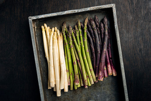 Overhead view of a metal tray full of freshly picked green, white and purple asparagus, photographed against a rustic wooden background. Colour, horizontal with some copy space.