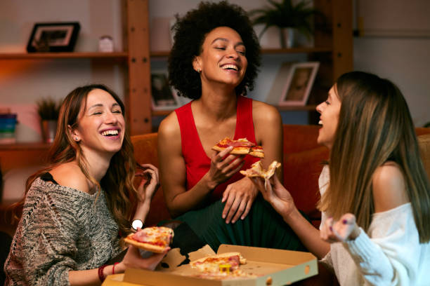 A multicultural group of cheerful young women is eating pizza while sitting in the living room. stock photo