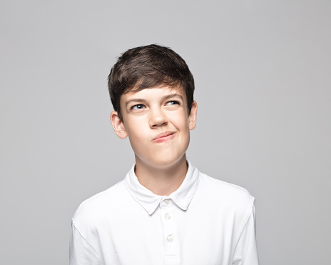 Confused teenage boy in t-shirt making face while standing against gray background.