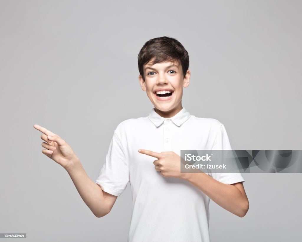 Excited teenage boy pointing against gray background Portrait of excited teenage boy pointing away while standing against gray background. Excitement Stock Photo