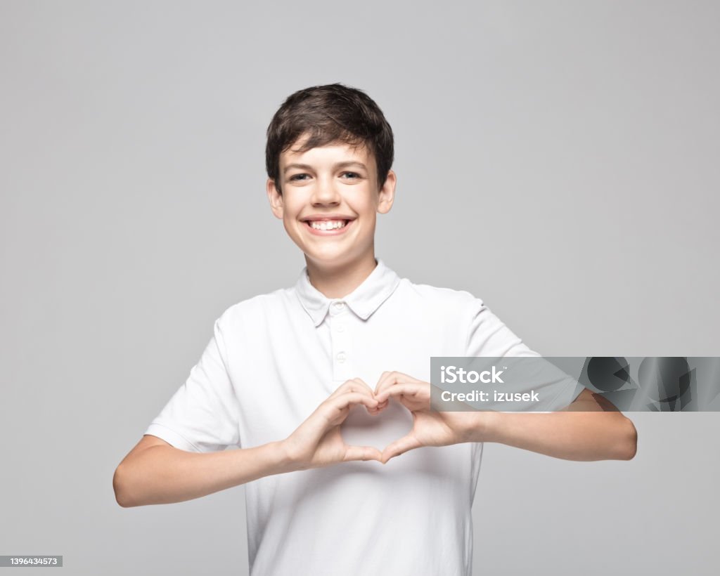 Happy teenage boy gesturing heart shape Portrait of happy teenage boy gesturing heart shape while standing against gray background. Heart Shape Stock Photo