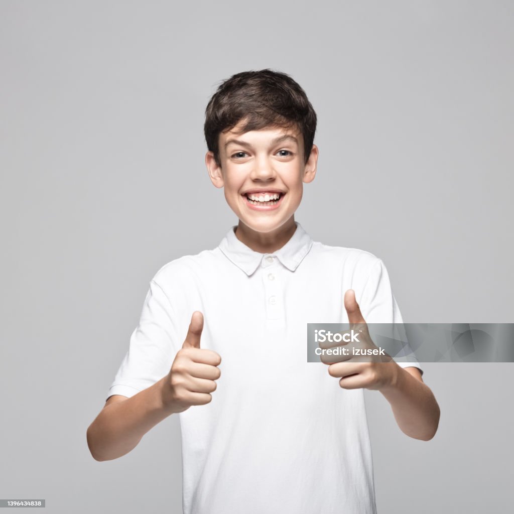 Portrait of happy boy gesturing thumbs up Cheerful teenage boy wearing t-shirt gesturing thumbs up while standing against gray background. Teenager Stock Photo