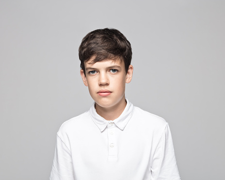 Portrait of sad teenage boy wearing white t-shirt standing against gray background.