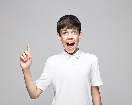 Surprised teenage boy with mouth open pointing while standing against gray background.