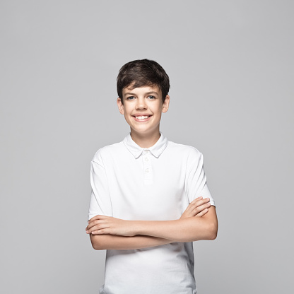 Portrait of happy teenage boy wearing white polo shirt standing with arms crossed against gray background.