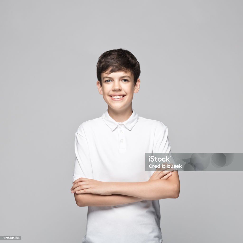 Teenage boy standing with arms crossed Portrait of happy teenage boy wearing white polo shirt standing with arms crossed against gray background. 14-15 Years Stock Photo