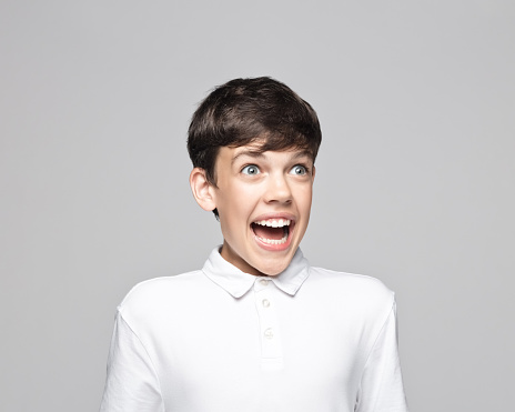 Excited teenage boy with mouth open looking away while standing against gray background.