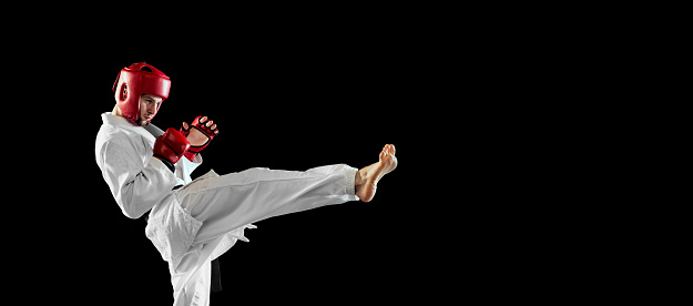 In motion. Male taekwondo fighter in white dobok, helmet and gloves training isolated over dark background. Concept of sport, education, skills, workout, health. Modern Korean martial art. Copy space for ad