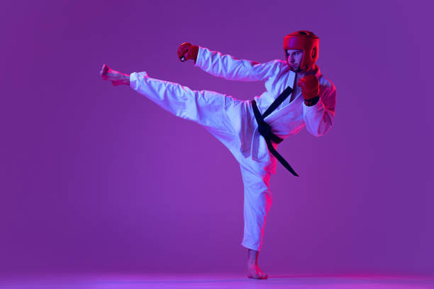 foto de estudio de un luchador masculino de taekwondo con uniforme deportivo en acción aislado sobre fondo púrpura en luz de neón. concepto de deporte, entrenamiento, competición, anuncio - purple belt fotografías e imágenes de stock