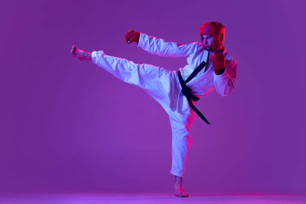 scatto in studio di combattente maschile di taekwondo in uniforme sportiva in azione isolato su sfondo viola in luce al neon. concetto di sport, allenamento, competizione, pubblicità - extreme sports karate sport exercising foto e immagini stock